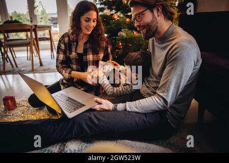 Junge mit seiner Mutter öffnet Weihnachtsgeschenke, während der Mann einen Videoanruf auf seinem Laptop führt. Familie zu Hause während einer Pandemie, die Weihnachten feiert. Stockfoto