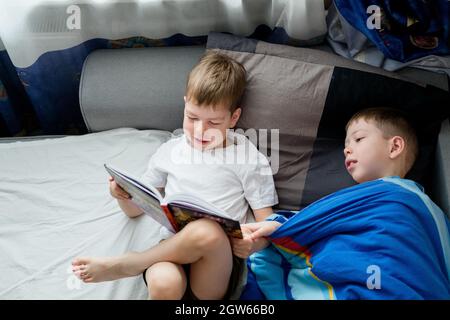 Der kleine Bruder liest ein Buch auf dem Bett für einen Ältesten. Freundschaft zwischen Brüdern. Liebe zum Lesen. Lerche und Eule unter den Menschen. 2 Jungen auf dem Bett Stockfoto