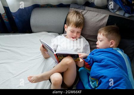 Der kleine Bruder liest ein Buch auf dem Bett für einen Ältesten. Freundschaft zwischen Brüdern. Liebe zum Lesen. Lerche und Eule unter den Menschen. 2 Jungen auf dem Bett Stockfoto