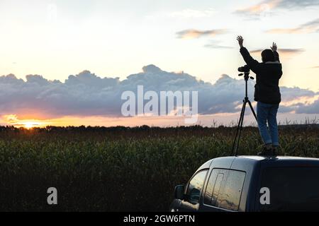 Ein Mädchen fotografiert den Sonnenuntergang von einem Stativ aus, das auf dem Dach eines Autos auf dem Feld steht. Stockfoto