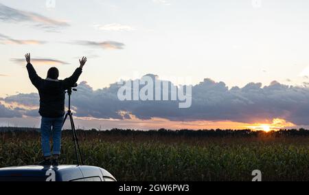 Ein Mädchen fotografiert den Sonnenuntergang von einem Stativ aus, das auf dem Dach eines Autos auf dem Feld steht. Stockfoto