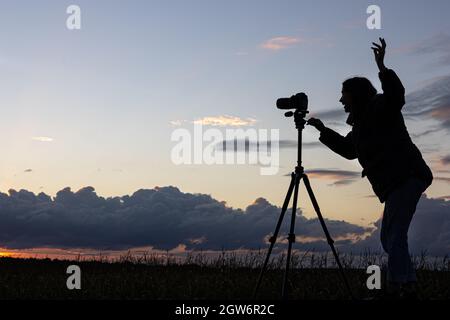 Ein Mädchen fotografiert den Sonnenuntergang von einem Stativ aus, das auf dem Dach eines Autos auf dem Feld steht und den Raum kopiert. Stockfoto