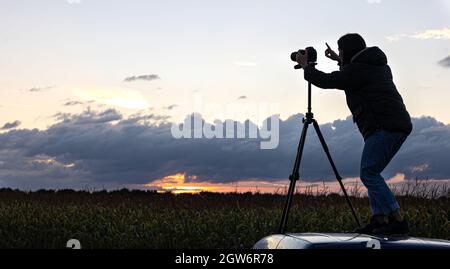 Ein Mädchen fotografiert den Sonnenuntergang von einem Stativ aus, das auf dem Dach eines Autos auf dem Feld steht und den Raum kopiert. Stockfoto