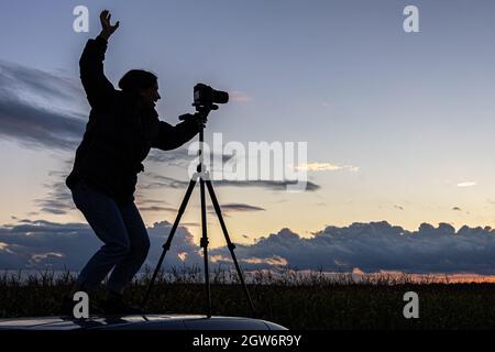 Ein Mädchen fotografiert den Sonnenuntergang von einem Stativ aus, das auf dem Dach eines Autos auf dem Feld steht und den Raum kopiert. Stockfoto