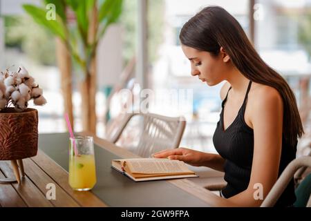 Junge hübsche Frau sitzt und trinkt eine Glaslimonade, während sie ein Buch liest. Hochwertige Fotos Stockfoto