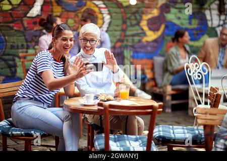 Zwei kaukasische Frauen, unterschiedlichen Alters, sitzen im Café im Freien, benutzen Handy für die Online-Kommunikation, sprechen mit jemandem, winken. Stockfoto