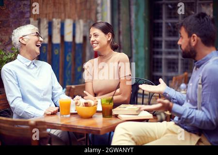 Zwei Frauen, junge Erwachsene und ältere Menschen, die sich über den bärtigen Kerl lustig machen, einander schauen, lachen, lächeln. Öffentlicher Ort, Café im Freien Stockfoto