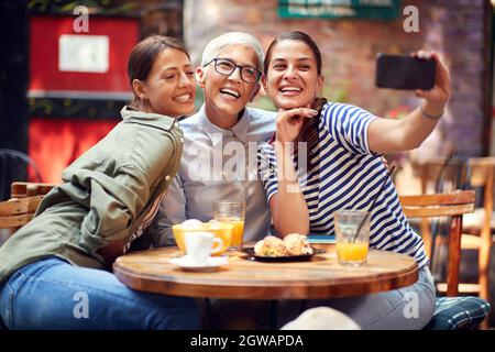 Zwei junge, schöne kaukasische Erwachsene Frauen, die mit einer älteren Frau ein Selfie am Handy machen, sitzen, lächeln, Stockfoto