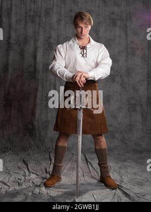 Ein junger Mann in einem Lederkilt und einer weißen Schnürbluse. Ein schottischer Ritter mit einem zweihändigen Schwert. Foto in voller Länge im Studio auf einem grauen Hintergro Stockfoto