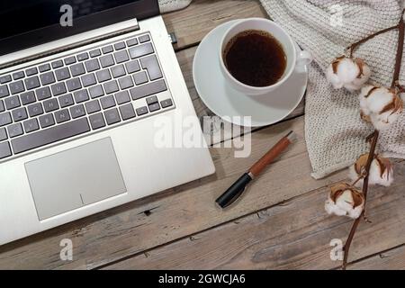Teil einer Laptop-Computer-Tastatur, Kaffeetasse und Stift auf einem rustikalen Holztisch mit Baumwollzweig und einem weißen Schal, Home-Office-Geschäft im Herbst, Stockfoto