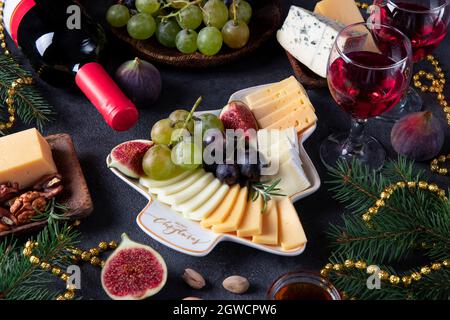 Verschiedene Käsesorten und Früchte, serviert auf einem Teller als Weihnachtsbaum, auf dunkelgrauem Hintergrund mit zwei Gläsern Wein. Silvesterparty-Snack Stockfoto