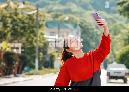Videokommunikation, Bloggen. Eine junge Frau posiert auf einer Telefonkamera, nimmt ein Selfie auf oder kommuniziert via Video-Link. Im Freien. Konzept der modernen Technologie Stockfoto