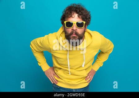 Überrascht Casual bärtiger Kerl, der die Hände an den Hüften hält und ein schockiertes Gesicht macht, während er vor blauem Hintergrund im Studio steht Stockfoto