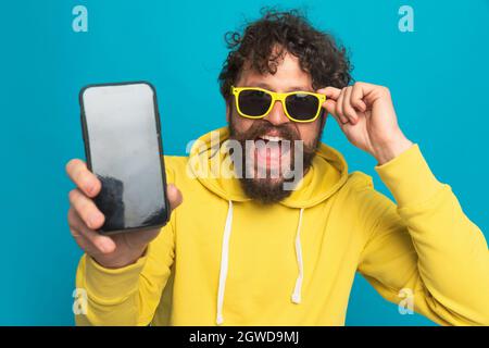 Enthusiastischer bärtiger Mann mit gelbem Kapuzenpullover, schwarzem leerem Bildschirm und fixierenden Sonnenbrillen, lächelnd und lachend auf blauem Hintergrund im Studio Stockfoto