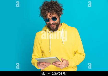 Cooler bärtiger Mann mit lockigen Haaren, der im Studio auf blauem Hintergrund steht und lächelt, Nachrichten liest und posiert Stockfoto