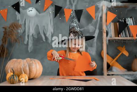 Kaukasisches Mädchen in einem Hexenkostüm sitzt zu Hause am Tisch und bereitet Kunsthandwerk für den Halloween-Urlaub vor. Stockfoto