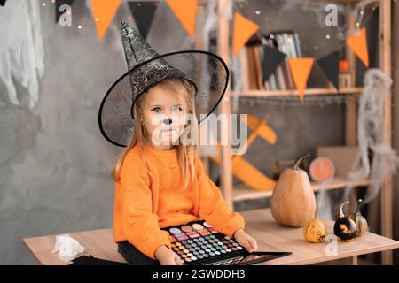 Portrait eines kleinen Mädchens in einem Halloween Kostüm, das am Tisch sitzt, mit einer Palette von Lidschatten für Make-up in einem Hexenhut. Urlaub am 31. Oktober Stockfoto