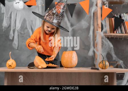 Ein fokussiertes kleines Mädchen bereitet eine Jack-o-Laterne für Halloween vor und nimmt Samen und Fruchtfleisch aus einem Kürbis. Kind in orangefarbenem Pullover und Hexenhut. Stockfoto