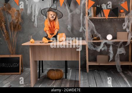 Ein Kind in einem Hexenhut bereitet eine Laterne für Halloween vor und nimmt Kürbiskerne und Fruchtfleisch heraus, während es auf einem Tisch in einem dekorierten Raum steht. Stockfoto