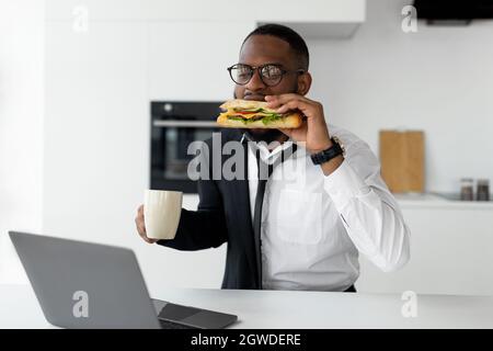 Moderner Geschäftiger Lebensstil. Porträt eines afrikanischen Mannes in einer Brille, der zu Hause in der Küche frühstückt, Anzug trägt und Kaffee trinkt, während er Sandw isst Stockfoto