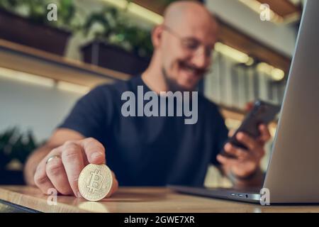 Der erfolgreiche kahle Geschäftsmann hält eine goldene Bitcoin-Münze in der Hand, während er das Smartphone vorführt. Stockfoto