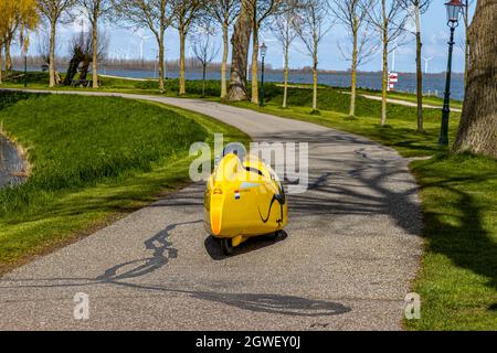 Medemblik, Noord-Holland, Niederlande. 14. April 2021. Rückansicht eines gelben Velomobils, das auf einem Pfad auf der Promenade zwischen Bäumen mit dem Fahrrad fährt Stockfoto