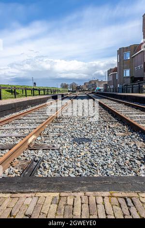 Zwei parallele Bahngleise mit ihren rostigen Schienen und Holzschwellern, zwischen grünem Gras und Gebäuden, Häusern und einem blauen Himmel im Hintergrund, sonnen sich Stockfoto