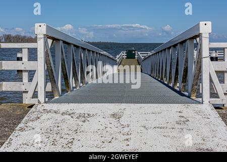 Gerader Fußweg mit einem niedrigeren Hang mit Horizont über dem IJsselmeer im verschwommenen Hintergrund, Holzbrücke, die zu einem Pier führt, sonniger Tag mit blauem Himmel Stockfoto