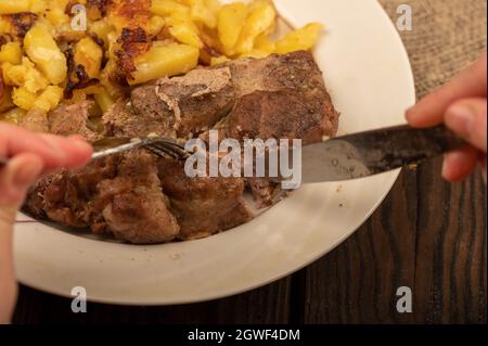Jemand schneidet mit einem Messer und einer Gabel ein Stück saftiges Braten auf einem Teller gebratener Kartoffeln. Nahaufnahme Selektiver Fokus Stockfoto