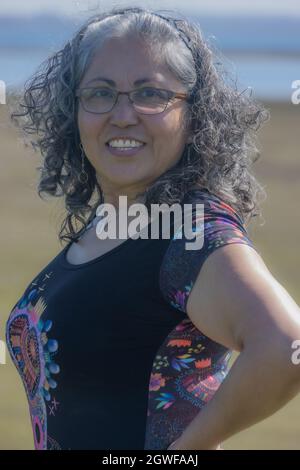 Fröhliche, reife Lateinerin mit einem breiten Lächeln und Blick auf die Kamera, vom Wind zerzauste graue, wellige Haare, gebräunte Haut, Brille, Stirnband, schwarzer Blou Stockfoto