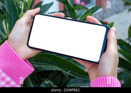 Mockup Bild von Nahaufnahme Frau Hand hält Handy mit leeren weißen Desktop-Bildschirm im Freien. Grüne Blätter im Hintergrund. Stockfoto