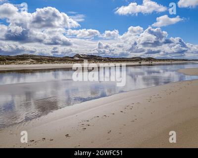 Wasser, das weiße Wolken auf dem Strandsand reflektiert, Menschen, die mit Dünen im Hintergrund wandern, wolkiger Tag mit reichlich Wolken am Himmel in Hargen aan Stockfoto