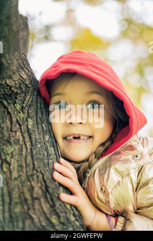 Ein Mädchen in einer goldenen Jacke und einer roten Kapuze sitzt auf einem Baum und lächelt zahnlos Stockfoto