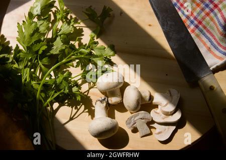 Flache Laie eines hölzernen Küchenbretts, auf dem einige Pilze geschnitten werden. Um uns herum sehen wir ein Messer und einige Petersilienzweige, mit Sonnenlicht, das thr Stockfoto