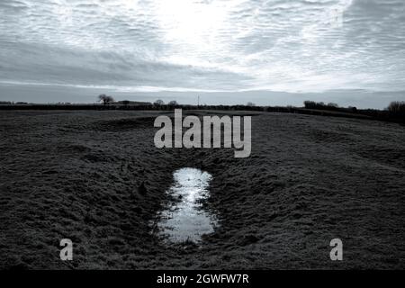 Im Vordergrund spiegelt ein Wasserfleck, der sich in einer Senke in einem Feld sammelt, einen gewaltigen dramatischen Himmel wider - Hemingford Abbots, Cambridgeshire Stockfoto