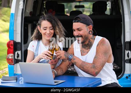 Junge tätowierte Paare lächeln, stoßen mit Bier in einem Laptop-Videoanruf an. Stockfoto