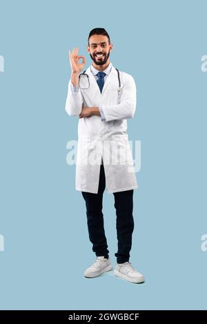 Fröhlicher Arabischer Arzt Gestikuliert Okay Zeichen Genehmigung Medizinische Behandlung Stehen Über Blauem Hintergrund Im Studio, Lächelnd Zur Kamera. Vertikal, Voll Stockfoto
