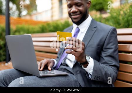 Junger afroamerikanischer Geschäftsmann mit Kreditkarte und Laptop, beim Online-Shopping auf der Bank im Freien sitzend, selektiver Fokus Stockfoto