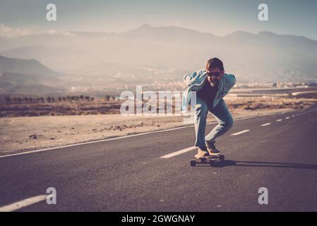 Reifer Mann in Sonnenbrille mit den Händen zurück auf seinem Longboard auf Landstraße mit Bergen im Hintergrund fahren. Man Skating oder Longboarding auf Mountain Ro Stockfoto