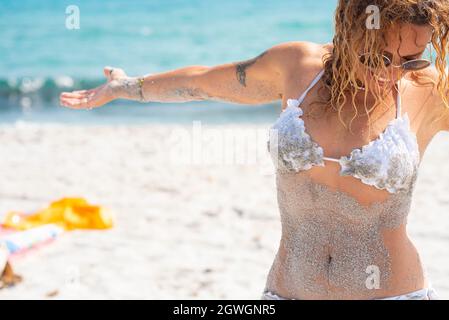 Glückliche reife Frau in weißem Bikini, die eine mit Sand bedeckte Sonnenbrille am Bauch trägt, während sie am Strand Arm ausgestreckt steht. Sorglose Frau in Bikini Co Stockfoto
