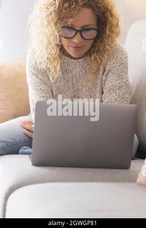 Schöne reife Frau mit lockigen Haaren, die eine Brille trägt und auf der Couch sitzt, während sie zu Hause einen Laptop benutzt. Frau, die mithilfe von Lapto Social-Media-Inhalten surft Stockfoto