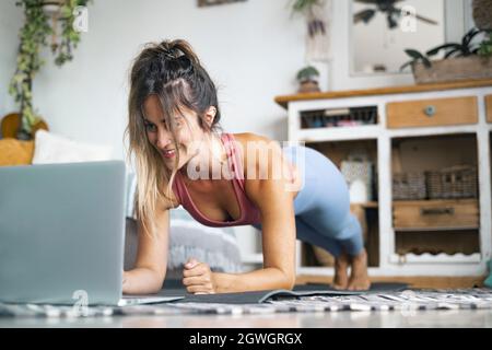 Schöne junge Frau, die Plank-Übung macht, während sie auf den Laptop auf dem Boden schaut. Frau in Sportkleidung, die zu Hause Pilates macht. Frau in Sportbekleidung Sport treiben Stockfoto