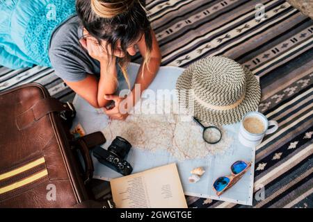 Blick aus der Perspektive einer jungen Frau, die mit einer Kaffeetasse auf dem Teppich liegt und den Atlas betrachtet. Junge Frau, die für die Lage auf der Karte mit Lupe auf Stockfoto