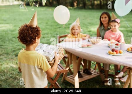 Porträt eines lockigen Jungen, der während einer Geburtstagsfeier im Freien mit Freunden im Sommer Geschenke trägt, Kopierfläche Stockfoto