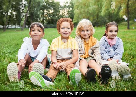In voller Länge Porträt einer Gruppe von Kindern, die im Park auf Gras sitzen und die Kamera betrachten Stockfoto