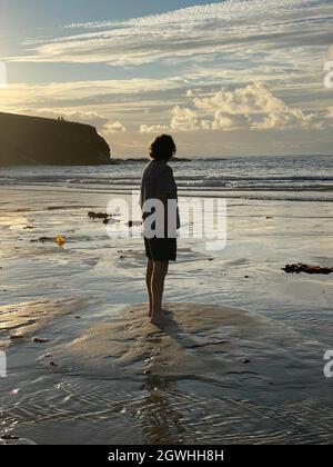 Volle Rückenansicht eines reifen Mannes, der am späten Nachmittag am Strand steht, als die Sonne untergeht. Mit Shorts und T-Shirt zum Horizont schauend. Stockfoto