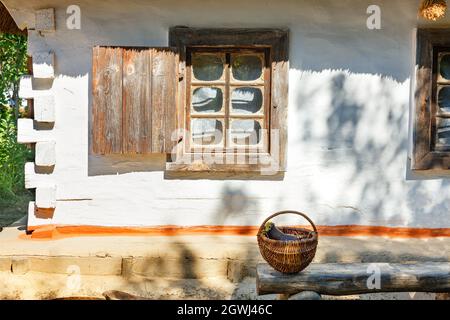 Ein altes Fenster mit hölzernen Fensterläden einer ukrainischen ländlichen Hütte unter einem Strohdach wird von den Sonnenstrahlen beleuchtet. Stockfoto