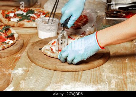 Nahaufnahme der Hände eines kaukasischen Mannes mit blauen Latexhandschuhen, der eine gekochte Pizza auf einem hölzernen Küchenbrett schneidet. Nahaufnahme, Kopierbereich. Stockfoto