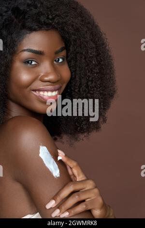 Lächelndes, fröhliches junges afro-amerikanisches Mädchen, das Creme auf den Körper aufgibt. Hautpflegekonzept. Stockfoto