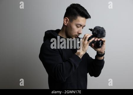 Ein Porträt eines hübschen ostasiatischen Kameramanns, der eine Kamera an einer grauen Wand testet Stockfoto